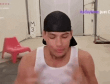a man wearing a white tank top and a black hat is standing in a room with a chair .
