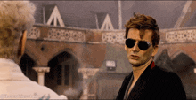 a man wearing sunglasses is talking to another man in front of a brick building