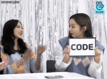 two young women are sitting at a table holding a sign that says code .