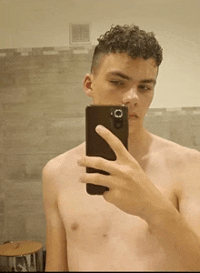 a young man without a shirt is taking a selfie with his cell phone .