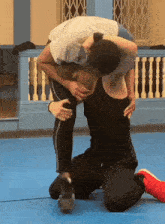 two men are wrestling on a blue mat and one of them is wearing a white shirt that says ' x ' on it
