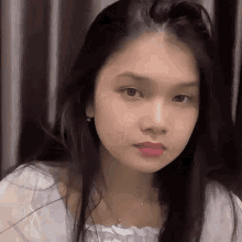 a young woman with long black hair is wearing a white top and a necklace and looking at the camera .