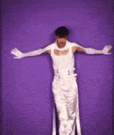 a man in a white tank top and white pants is standing in front of a purple background holding a knife .