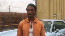 a man in an orange jacket stands in front of a blue car with betx written on the bottom