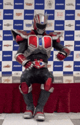 a man in a red and black costume is sitting in front of a blue and white checkered wall with the number 110