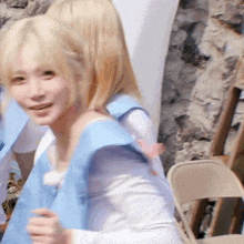a girl with blonde hair is wearing a blue vest and smiling while standing next to a chair .
