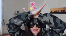 a woman wearing sunglasses and a camouflage jacket makes a peace sign with her hands