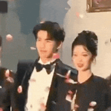 a man in a tuxedo and bow tie is standing next to a woman in a black dress .