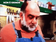 a bald man with a beard wearing overalls and an orange shirt is standing in front of a shelf with knives .