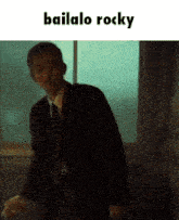 a man in a suit and tie is standing in front of a window with the words bailalo rocky written above him .