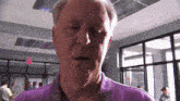 a man in a purple shirt stands in a hallway with a red exit sign in the background