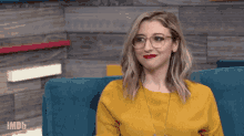 a woman wearing glasses and a yellow sweater sits on a blue couch with imdb written on the bottom right