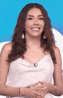 a woman wearing a white dress and earrings is smiling while standing in front of a blue background .