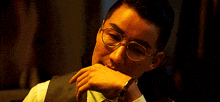 a man wearing glasses and a vest holds his hand to his mouth .