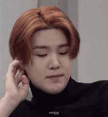 a young man with red hair is wearing a black turtleneck sweater and touching his ear .