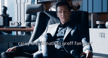 a man in a suit sits in a chair with the words casey the johnathan groff fan over written below him