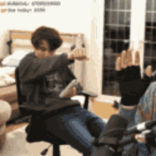 a man is sitting in a chair playing a video game while another person holds a remote control .