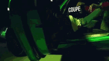 a man in a red jacket is sitting in a green car with the word coupe on his shirt .