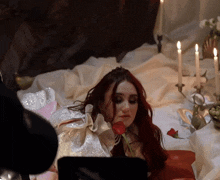a woman with red hair is laying on a bed with candles in the background