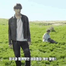 a man in a leather jacket is standing in a grassy field while another man sits on the grass .