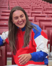 a woman in a red , white , and blue jacket is sitting in a stadium laughing .