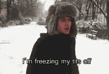 a man in a fur hat is standing in the snow and says i 'm freezing my tits off .