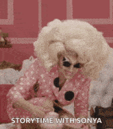 a drag queen is sitting on a bed holding a teapot and a book .