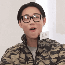 a young man wearing glasses and a camouflage jacket has a piece of paper on his neck that says ' highest '