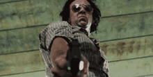 a man wearing sunglasses holds a gun in front of a wooden wall