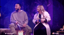 a man and a woman are sitting next to each other in front of a fire with the nbc logo in the background