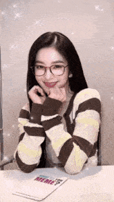 a woman wearing glasses is sitting at a table with a notepad that says " what do you meme ? " on it