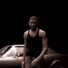 a man in a black tank top is dancing in front of a silver car