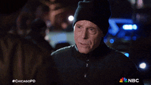 a man wearing a black hat is talking to another man in front of a police car with nbc written on the bottom right