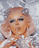 a close up of a drag queen laying on a bed with her finger to her mouth