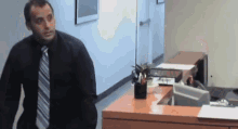 a man in a black shirt and tie is walking towards a desk