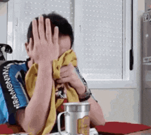 a man is covering his face with a towel while sitting at a table with a cup of coffee .