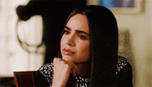 a woman with long dark hair is sitting at a table with her hand on her chin and looking to the side .