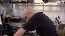 a bald man in a black shirt is cooking in a kitchen and says " arriba "