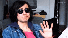 a young man wearing sunglasses and a denim jacket is waving his hand
