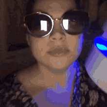 a woman wearing sunglasses looks at the camera with a blue light behind her