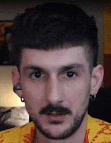 a man with a beard and earrings is wearing a yellow shirt and looking at the camera .
