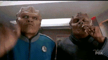 two aliens are standing next to each other in a room and one is smoking a cigarette .