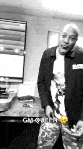 a man in a black jacket is standing in front of a desk with the words gm-queen on the bottom
