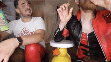 a man wearing a red leather jacket is smoking a cigarette next to a man wearing a white shirt that says pure passion