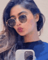 a woman wearing round sunglasses is taking a selfie with her phone