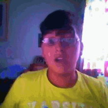 a young boy wearing glasses and a yellow shirt is talking to the camera while standing in front of a window .