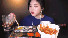a woman wearing ear buds is eating a tray of food with chopsticks
