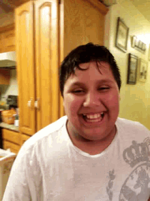 a boy wearing a white shirt with a crown on it is smiling in a kitchen