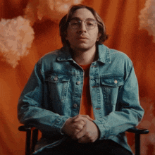 a man wearing a denim jacket and glasses sits with his hands folded