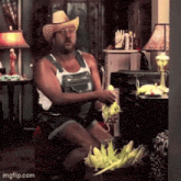 a man wearing overalls and a cowboy hat is sitting in a living room holding corn